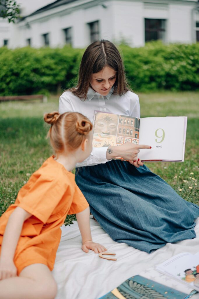 A woman reads a book to a child on a picnic blanket outdoors, promoting learning and bonding.